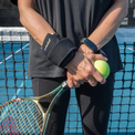 A close-up of a person on a tennis court wearing the VibraCool Easyfit on one wrist and holding a tennis racket and tennis ball. The brace has a white logo that reads 'TENSER'.