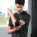 A man wearing a black athletic t-shirt demonstrates a VibraCool Easyfit for elbow support brace on his forearm in an indoor setting