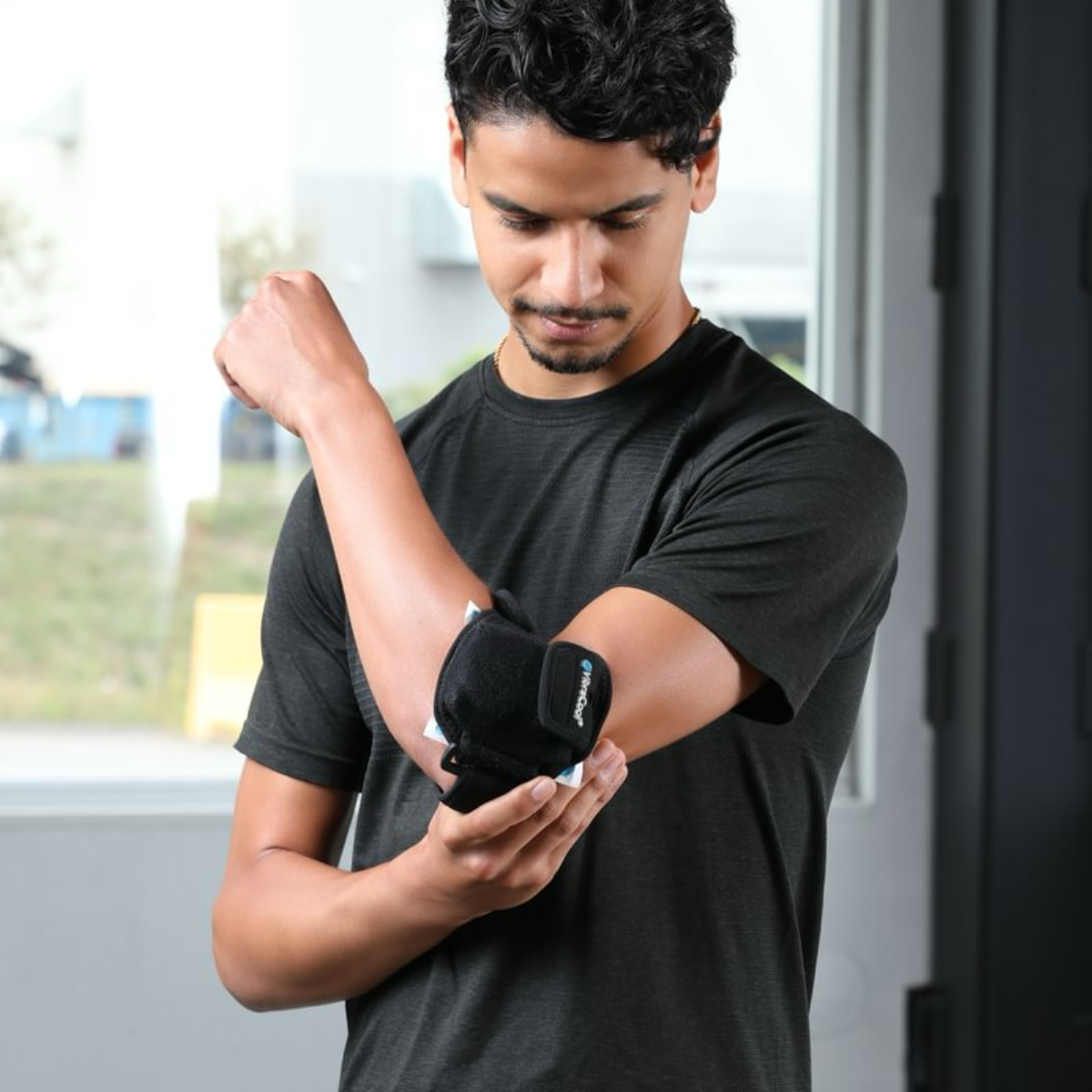 A man wearing a black athletic t-shirt demonstrates a VibraCool Easyfit for elbow support brace on his forearm in an indoor setting