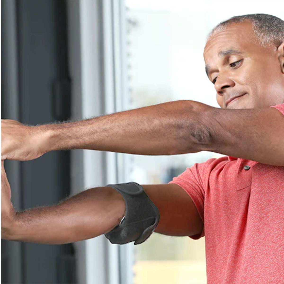 A man wearing a red polo shirt demonstrates the use of a VibraCool Easyfit on his elbow support brace on his forearm, with a window in the background.